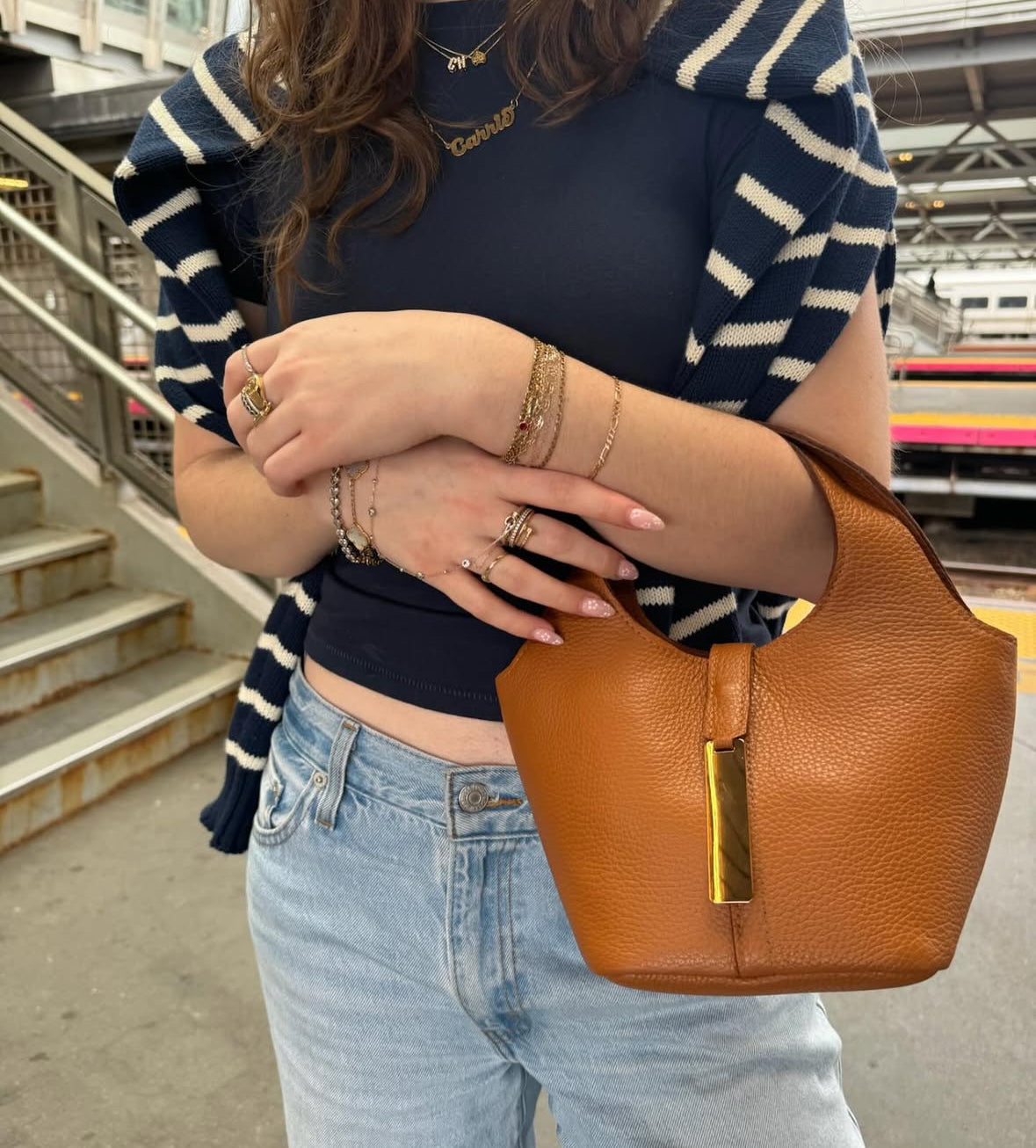 A person wearing a striped black and white top and light blue jeans, holding a brown leather crossbody bag with gold detailing.