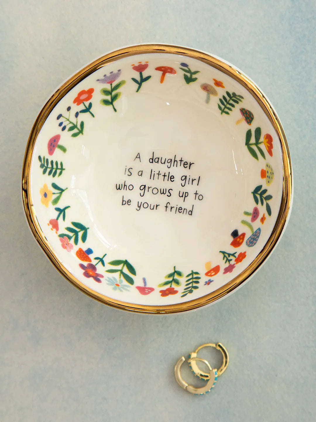 Decorative plate with floral designs and a quote, accompanied by a ring on a light blue background.